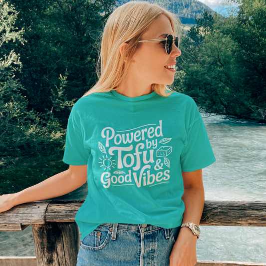 Woman standing on a bridge in a beautiful setting wearing a turquoise vegan t-shirt with the slogan 'powered by tofu and good vibes'