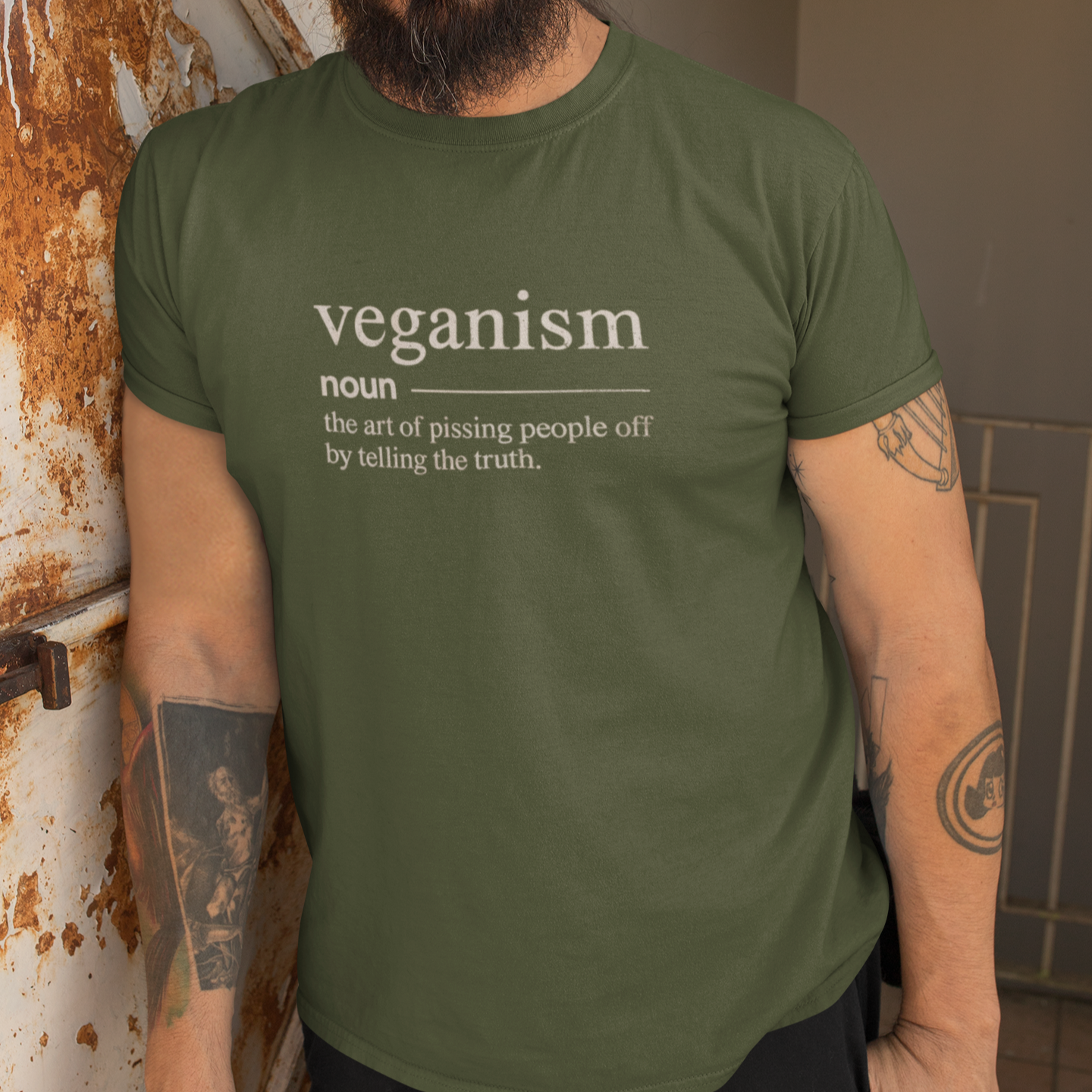 Bearded man wearing a khaki green funny vegan t shirt with the the dictionary definition 'veganism: the art of pissing people off by telling the truth'.
